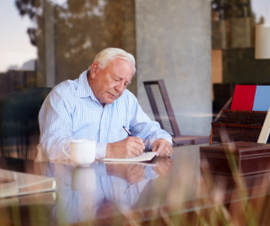 Starszy mężczyzna siedzi przy stole i pisze długopisem w notatniku.